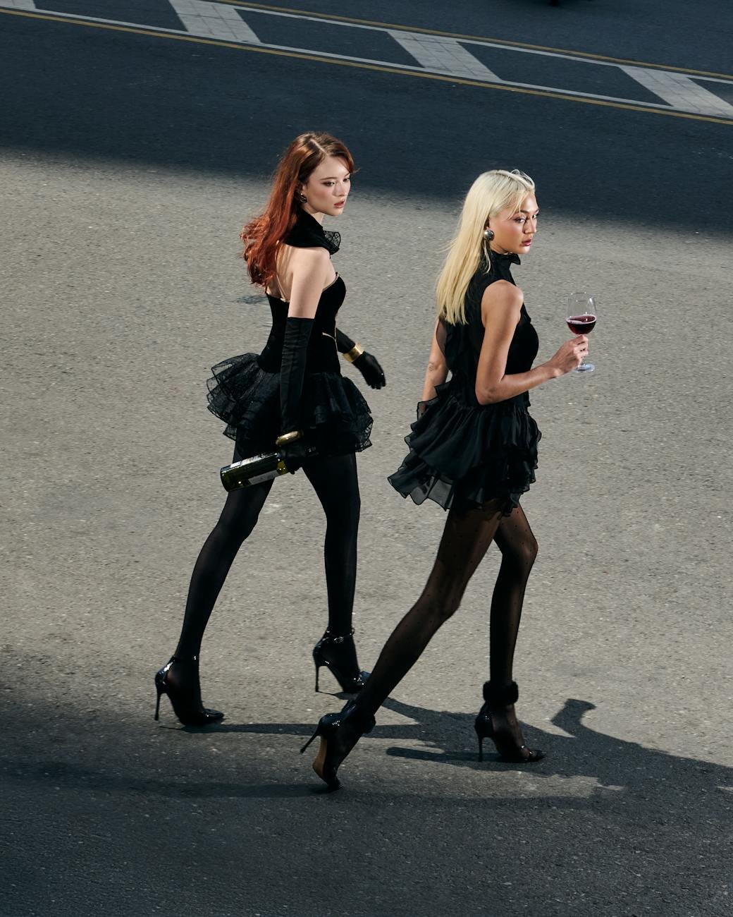 Two fashionable women in black dresses walking outdoors with wine bottles.